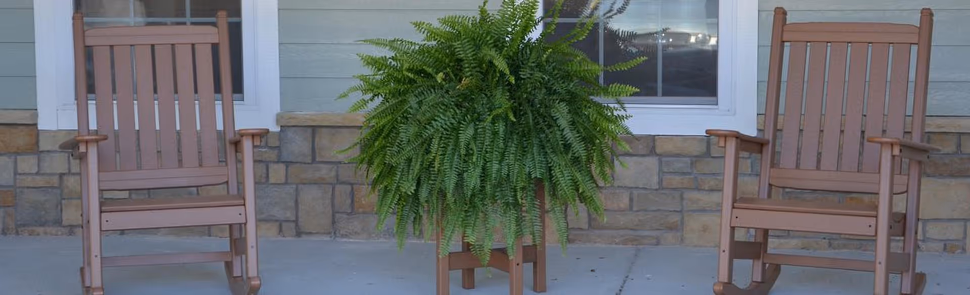 Two wooden rocking chairs on a porch with a large green fern plant in a wooden stand between them. The porch has a stone and siding exterior wall with a window behind the plant.