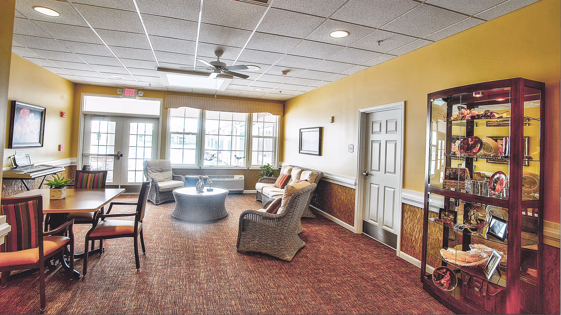 A cozy sitting area in a senior living facility with wicker chairs and a sofa around a round glass-top table. There is a wooden display cabinet with decorative items on the right, a table with chairs on the left, and large windows letting in natural light at the far end. The walls are painted yellow with a white chair rail and brown carpeted floor.