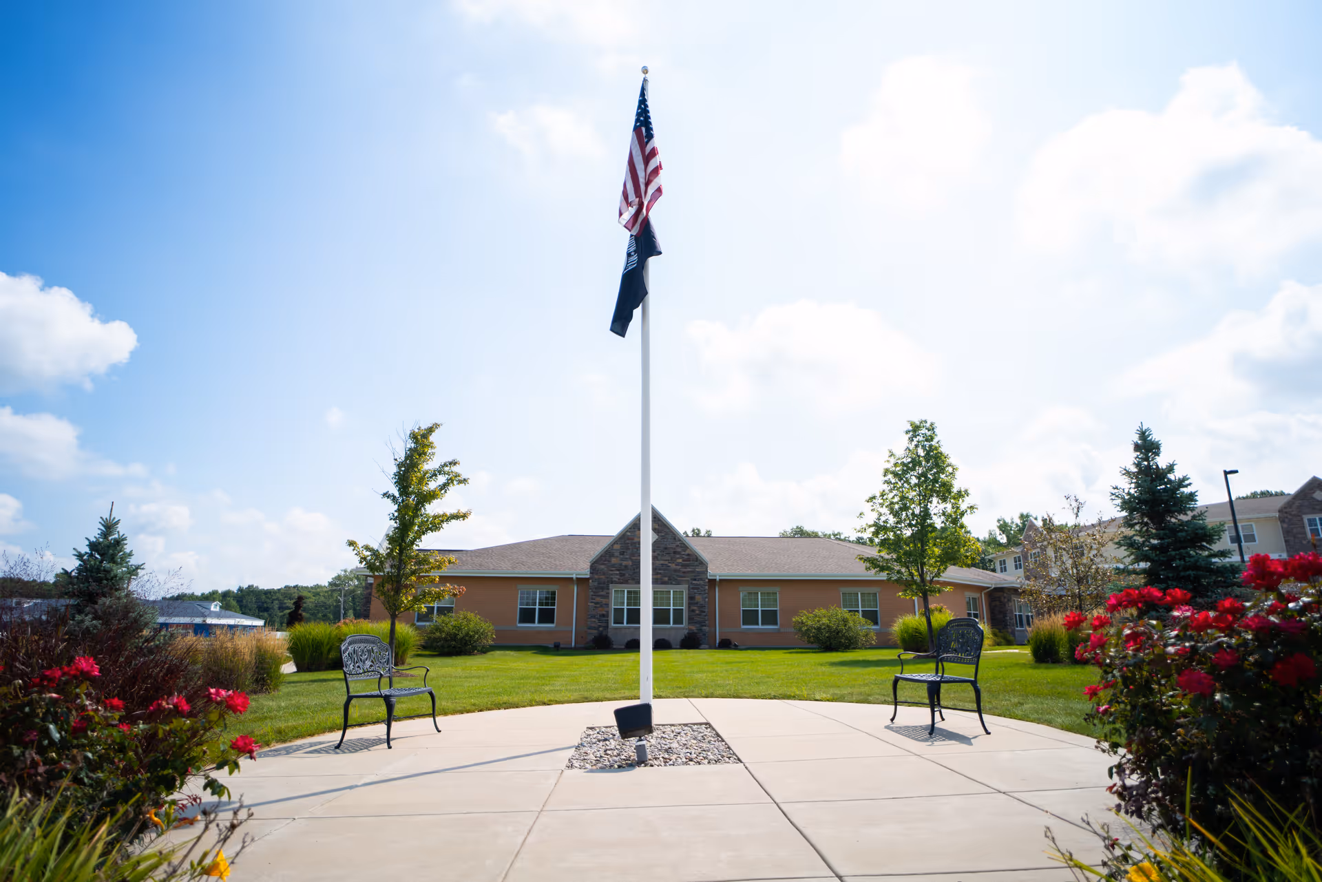 A flagpole with an American flag stands in a landscaped courtyard with benches and flowers in front of a one-story building.