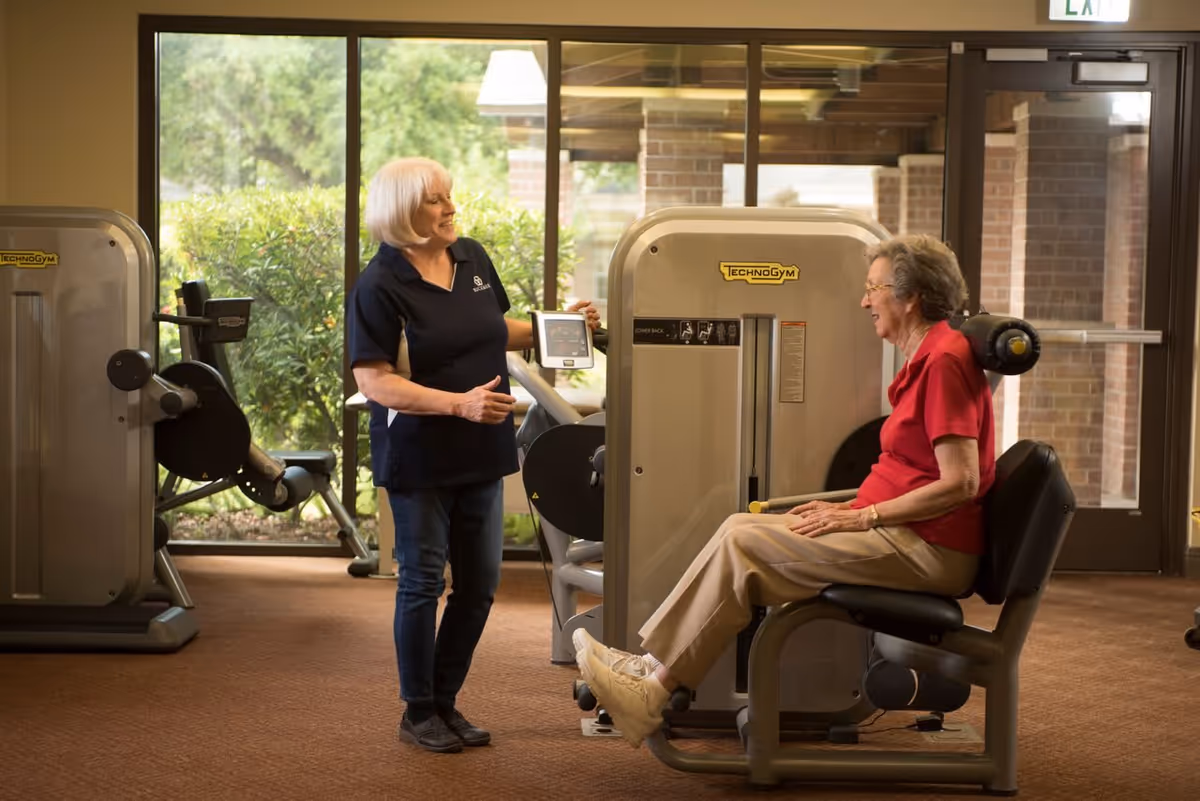 A staff member talks with an elderly woman seated on a leg exercise machine in a fitness room.