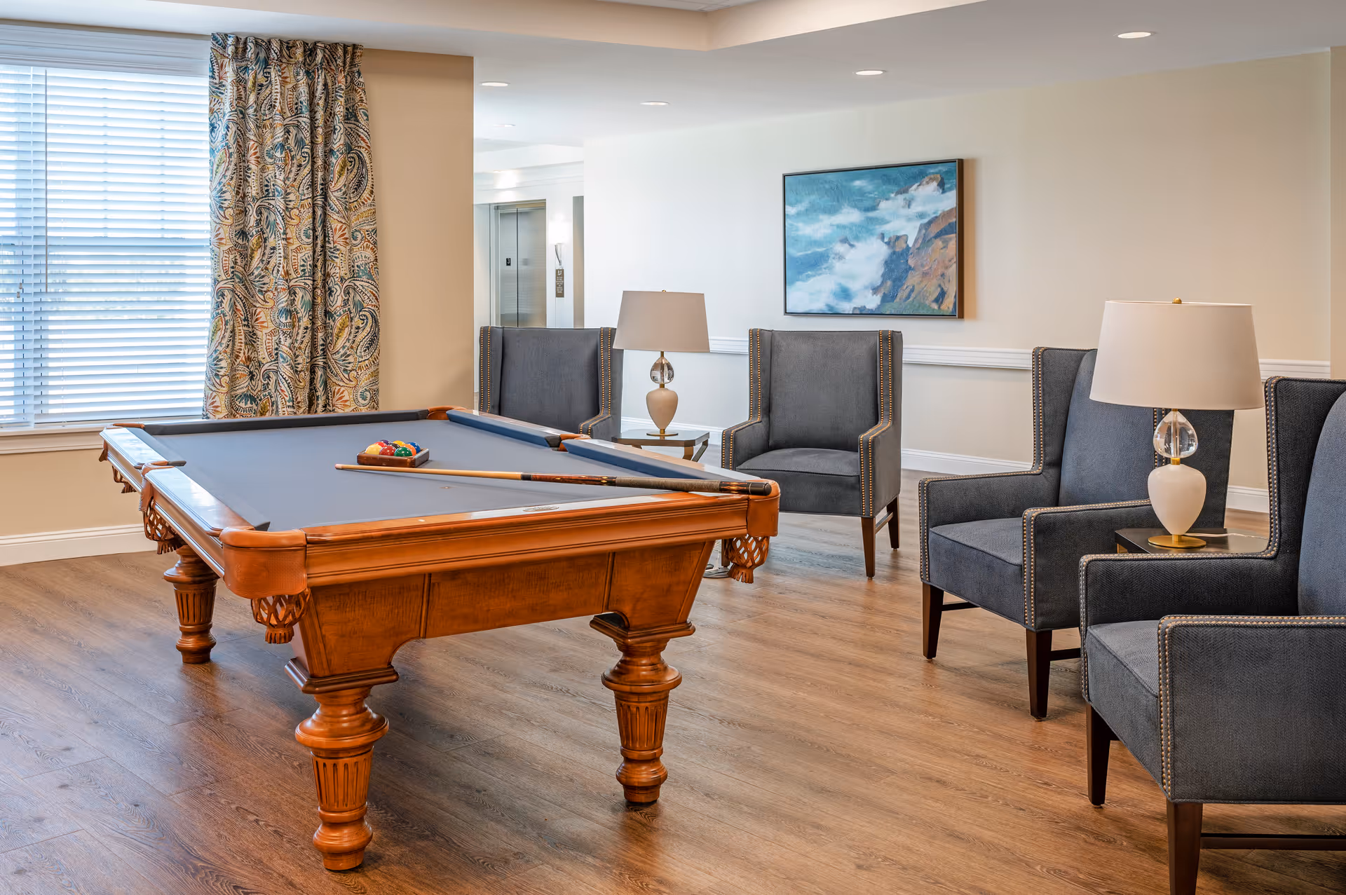 A well-lit room with a wooden pool table in the center, set up with billiard balls and cues. Surrounding the pool table are four gray upholstered armchairs with nailhead trim, two side tables each holding a lamp with a white shade. A large window with patterned curtains lets in natural light, and a painting of a coastal scene hangs on the beige wall.