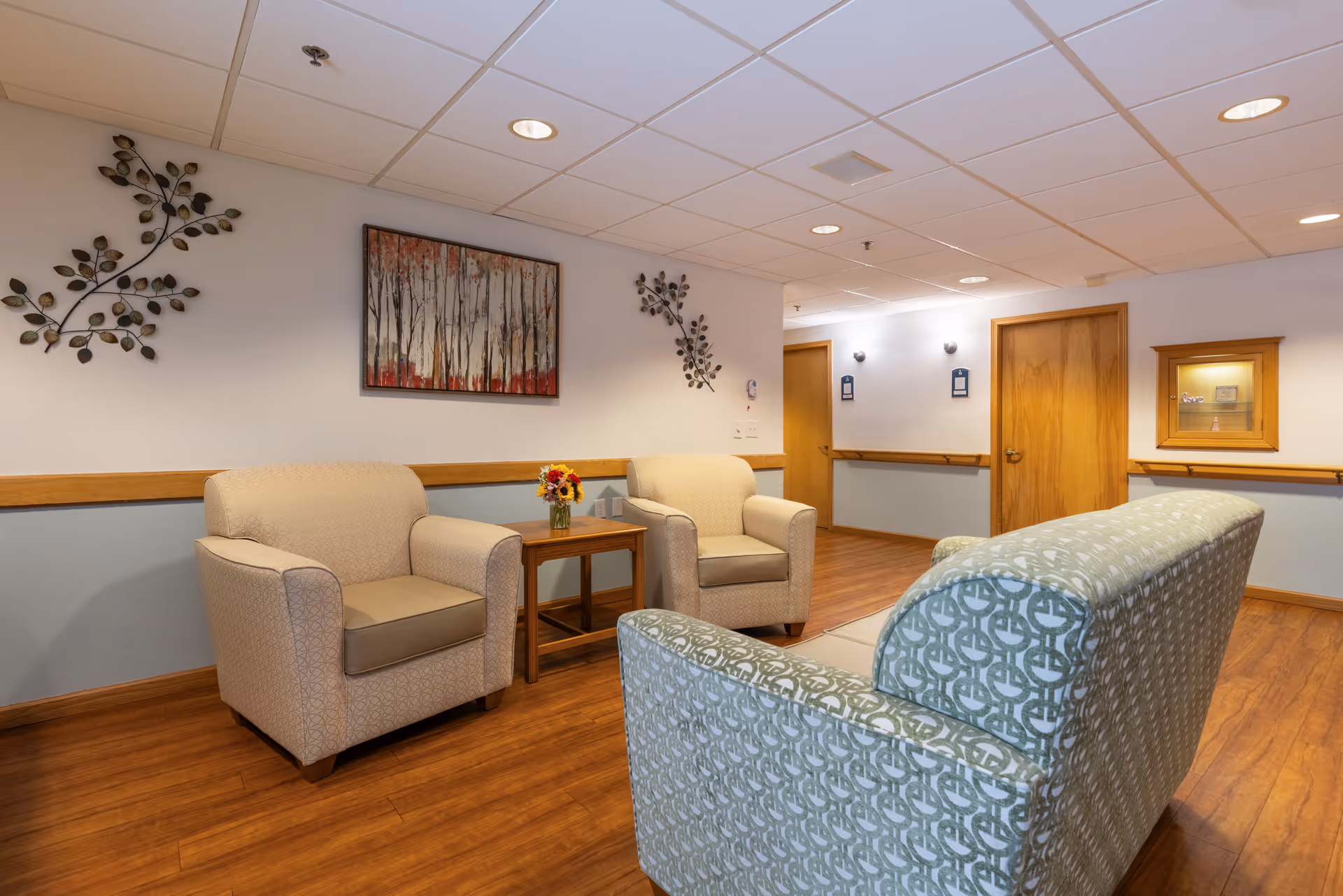 A cozy seating area in a senior living facility with two beige armchairs and a patterned sofa arranged around a small wooden table with a vase of flowers. The walls are decorated with metal leaf art and a framed painting of trees. The floor is wood, and there are wooden doors and handrails along the walls.