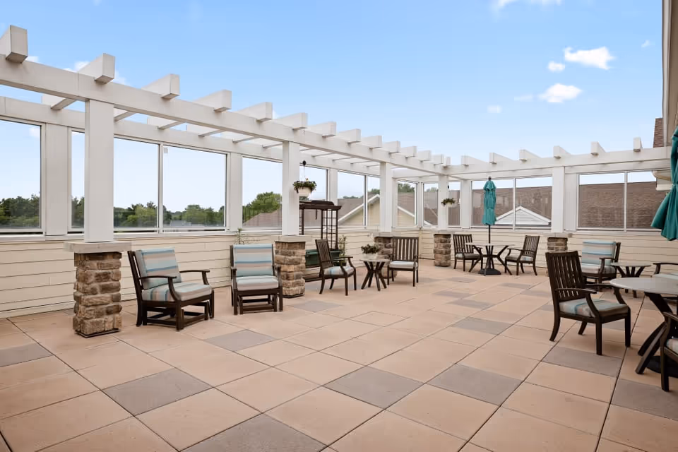 A spacious outdoor patio area with tiled flooring, several cushioned chairs, small tables, and a white pergola overhead. The patio is enclosed with glass panels and offers views of nearby rooftops and trees under a clear blue sky.