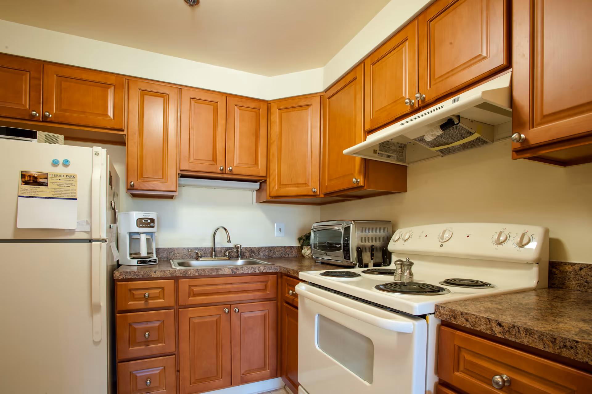 Compact kitchen with wooden cabinets, a white stove and refrigerator, a sink, coffee maker and toaster oven on granite-look counters.
