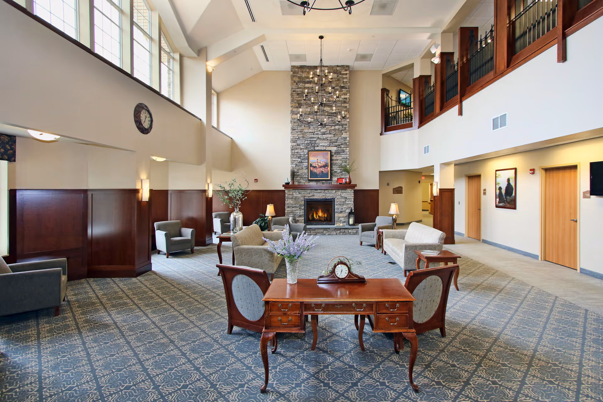 A spacious living room area with high ceilings and large windows letting in natural light. The room features a stone fireplace with a lit fire, surrounded by comfortable armchairs and sofas arranged for conversation. A wooden table with a clock and a vase of flowers is in the foreground. The walls are decorated with framed artwork, and there is a balcony railing visible on the upper level.