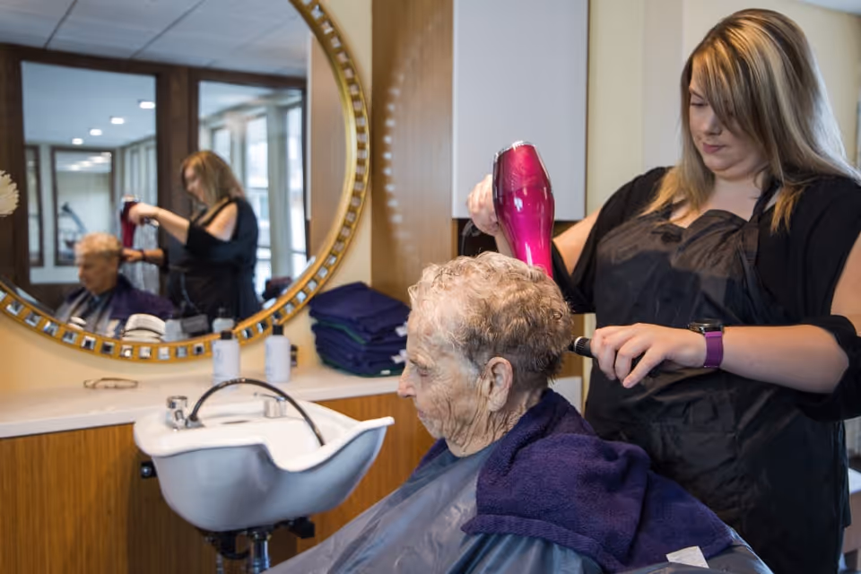 A hairdresser blow-drying the hair of an elderly woman seated in a salon chair in front of a large round mirror. The elderly woman is wearing a dark cape and a purple towel around her shoulders. The hairdresser is focused on styling the woman's short, curly hair.