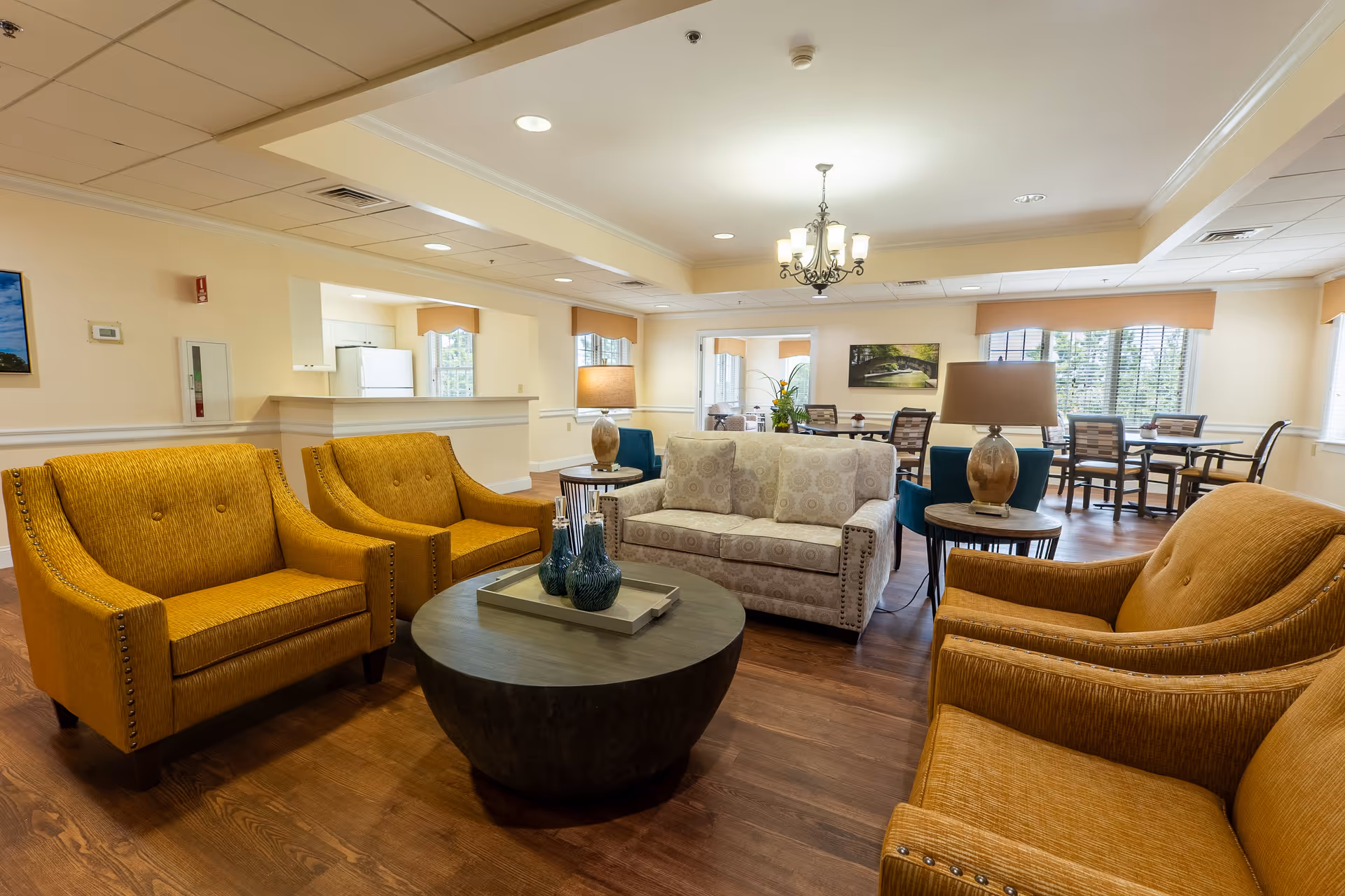 A spacious senior living common area with mustard yellow armchairs, a beige patterned sofa, a round dark wood coffee table with decorative vases, and side tables with lamps. In the background, there are dining tables and chairs near large windows with blinds, and a kitchen area with a white refrigerator is visible.