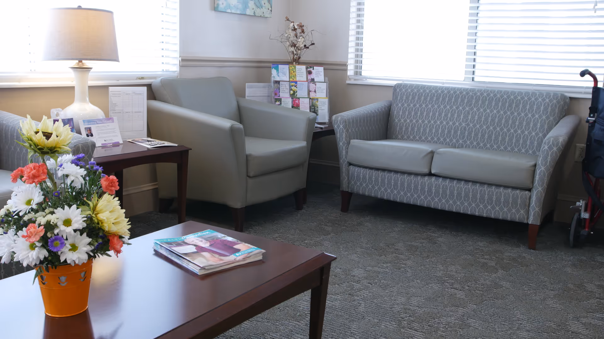 A cozy sitting area in a skilled nursing facility with a patterned loveseat, a gray armchair, a wooden coffee table with magazines, a side table with a lamp and informational brochures, and a colorful flower arrangement in an orange pot. A walker is partially visible on the right side near the window.