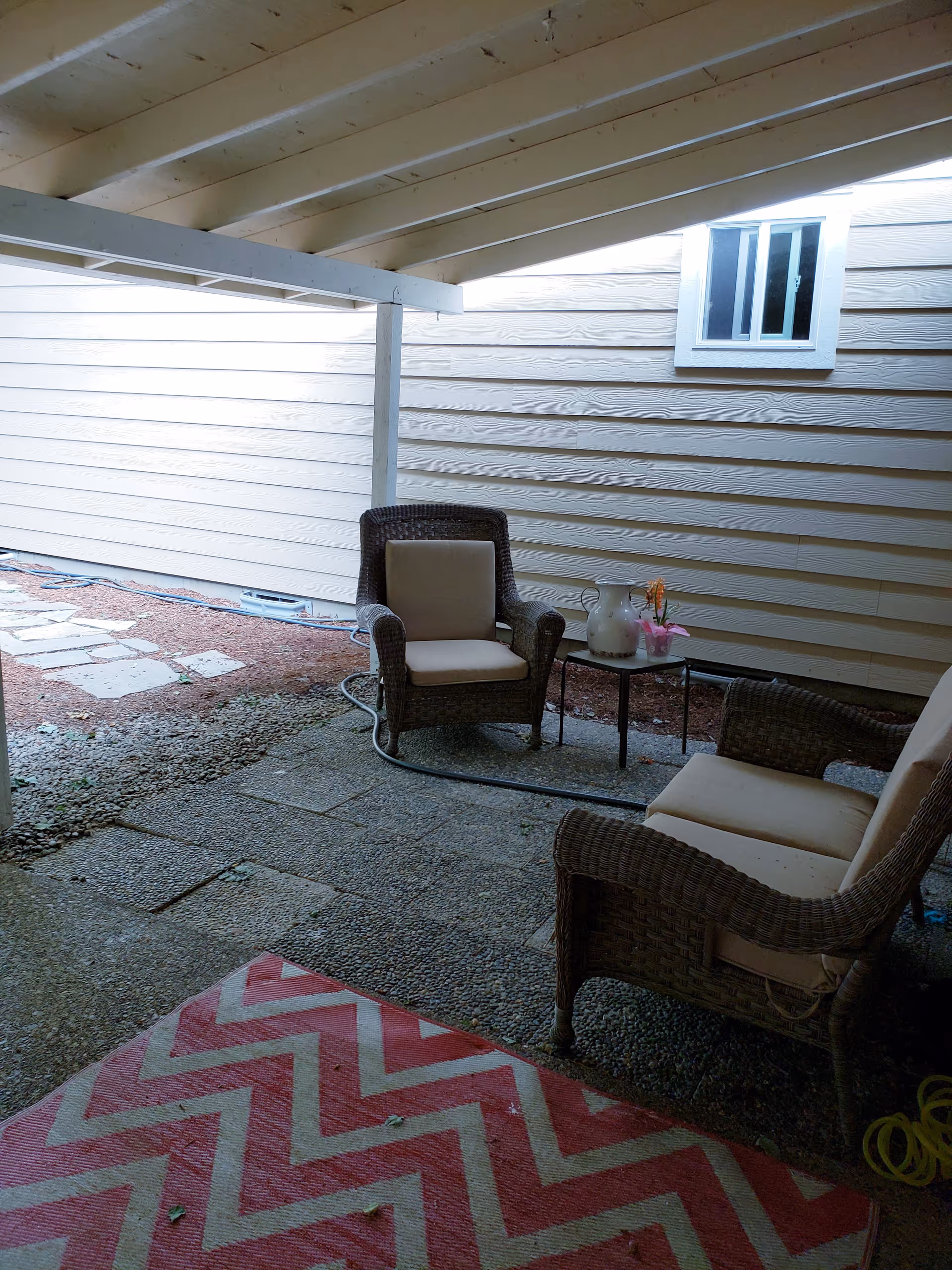 Covered outdoor patio area with two wicker chairs with beige cushions, a small table with a white pitcher and a small potted plant, a red and white zigzag patterned rug on the ground, and a beige siding wall with a small window.