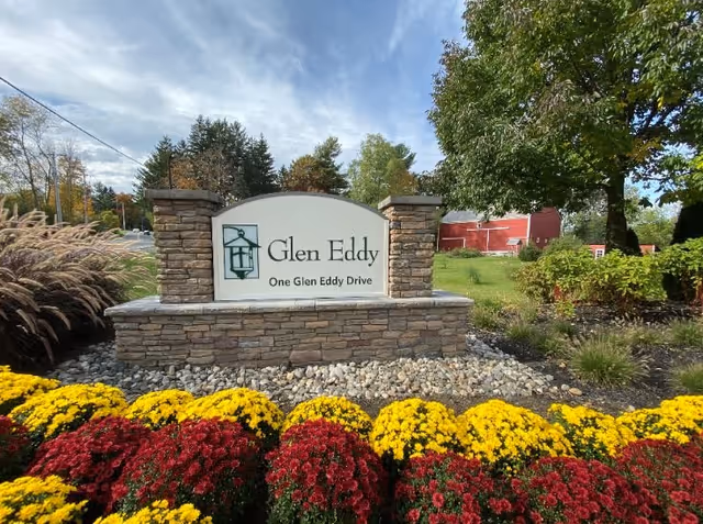 Stone sign for Glen Eddy at One Glen Eddy Drive surrounded by colorful red and yellow flowers, with trees and a red barn in the background under a partly cloudy sky.