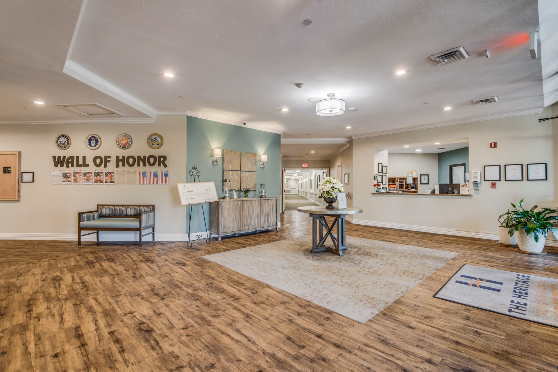 Lobby of a senior living facility with a central round table, reception desk, seating and a 'Wall of Honor' display on the wall.