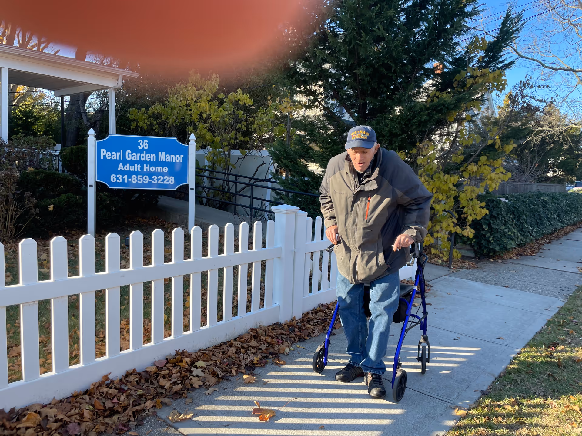 An elderly man using a walker approaches a sign for Pearl Garden Manor Adult Home.