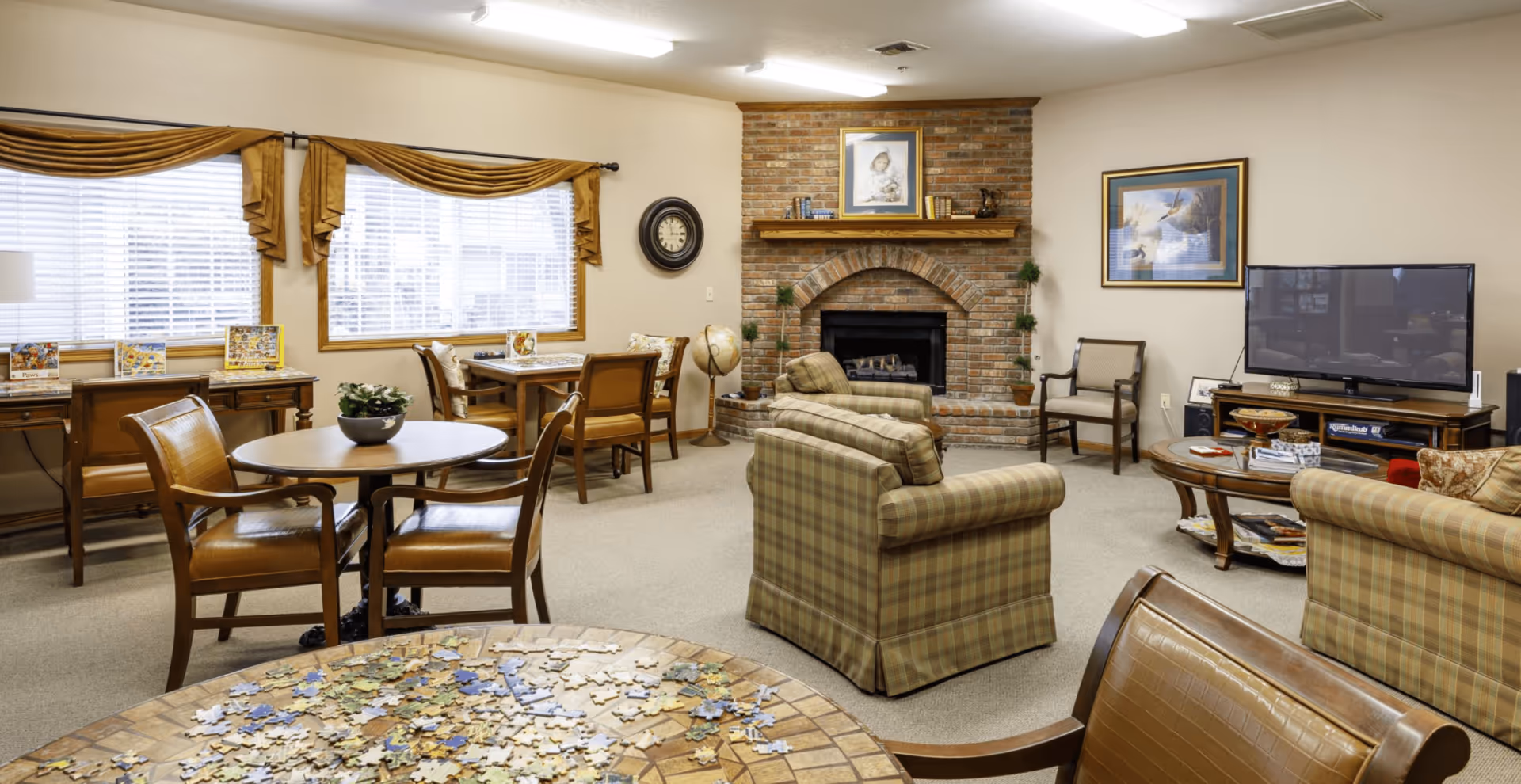 A cozy living room area in a senior living facility with plaid upholstered armchairs, a brick fireplace with a framed picture above it, a flat-screen TV on a wooden stand, multiple wooden tables and chairs, a globe, and windows with gold curtains letting in natural light. A table in the foreground has a partially completed jigsaw puzzle.
