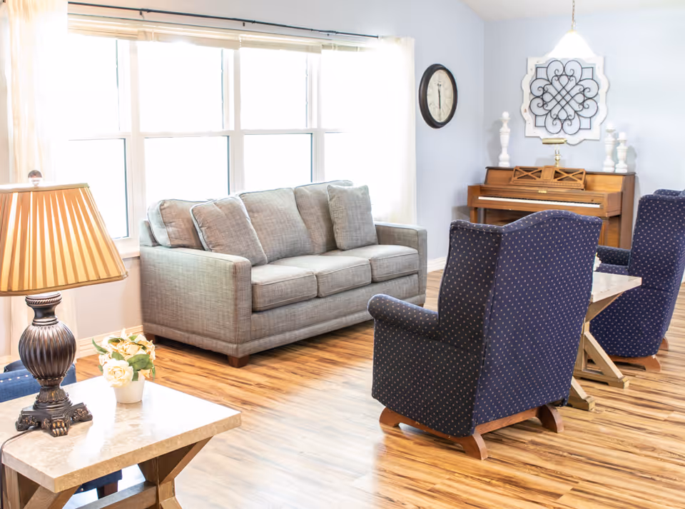 A bright living room with a large window covered by sheer curtains, a gray sofa with cushions, two blue patterned armchairs, a wooden piano against the wall, a round wall clock, decorative wall art, a table lamp on a side table, and a small flower arrangement on the table.