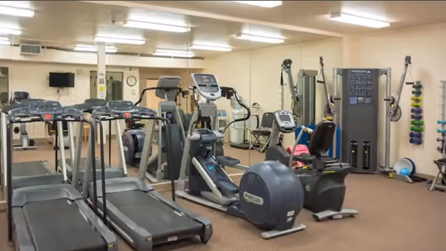 Interior view of a fitness room at Crista Shores featuring multiple exercise machines including treadmills, stationary bikes, elliptical trainers, and weight machines. The room has beige walls, bright overhead lighting, and a wall-mounted TV.