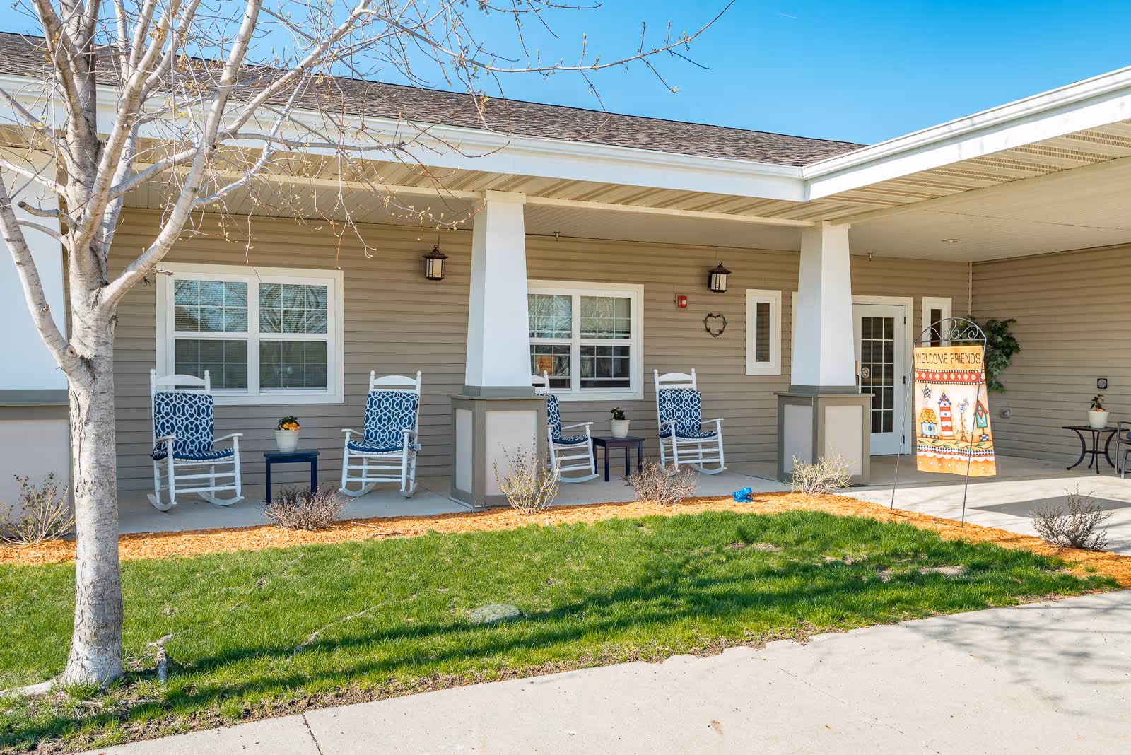 Covered front porch of Windsor Manor with rocking chairs, potted plants, and a welcome flag.
