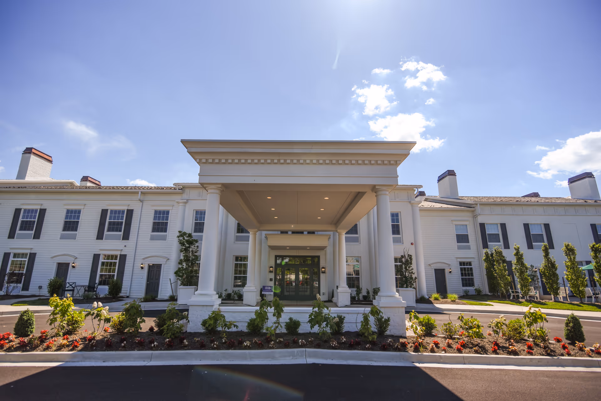 Front exterior view of a large white senior living facility building with a covered entrance supported by columns, landscaped garden beds with flowers and shrubs, and a clear blue sky with some clouds.