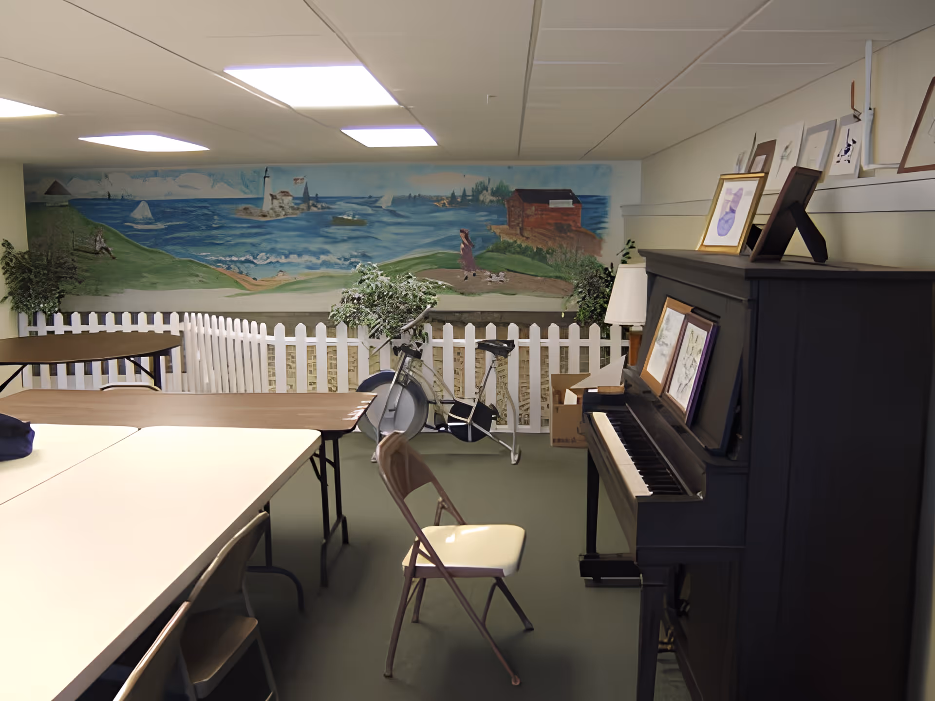 An interior room with tables and chairs arranged for group activities. A black upright piano with framed pictures on top is on the right side. In the background, there is a white picket fence with green plants and a stationary exercise bike. The back wall features a mural depicting a coastal scene with a lighthouse, sailboats, and a person walking along a path.