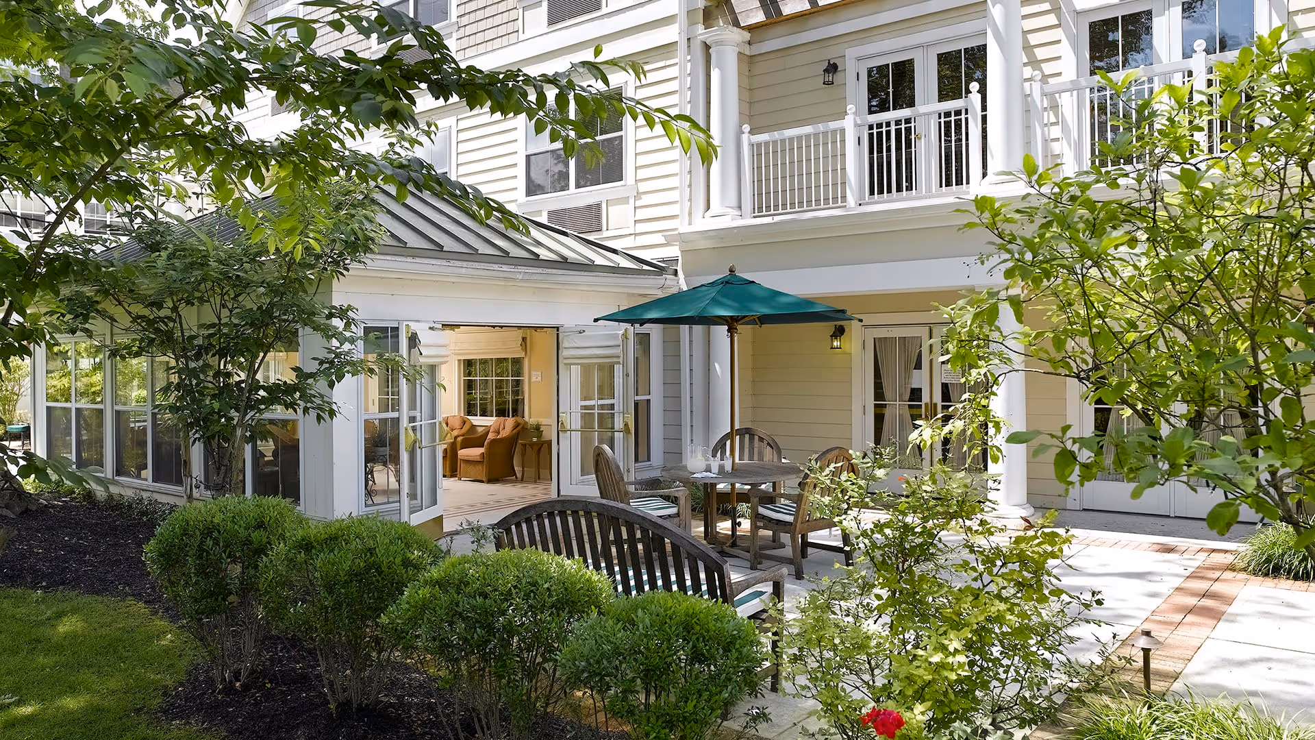 Outdoor patio area at a senior living facility with wooden chairs and tables, one table shaded by a green umbrella. The patio is surrounded by green bushes and trees, with a building featuring large windows and a balcony in the background.