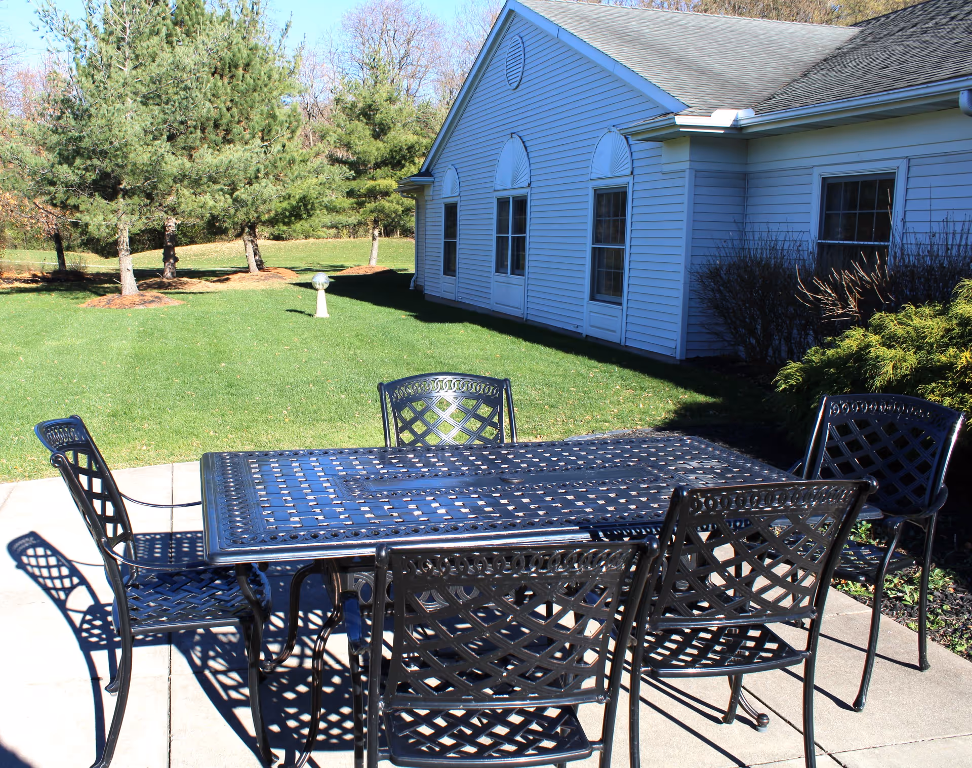 Outdoor patio area with a black metal table and six matching chairs on a concrete surface. The patio is adjacent to a white building with several windows. In the background, there is a well-maintained green lawn with trees and shrubs under a clear blue sky.