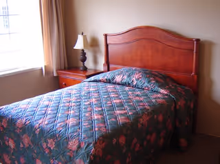 A bedroom with a wooden headboard bed covered with a floral patterned quilt. There is a wooden nightstand next to the bed with a decorative lamp on it. A window with beige curtains is visible on the left side of the room.