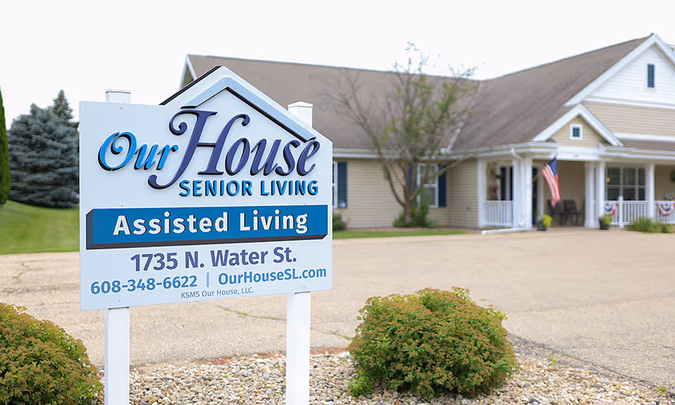 A white sign reading 'Our House Senior Living Assisted Living' stands in front of a single-story assisted living building with a porch and landscaping.