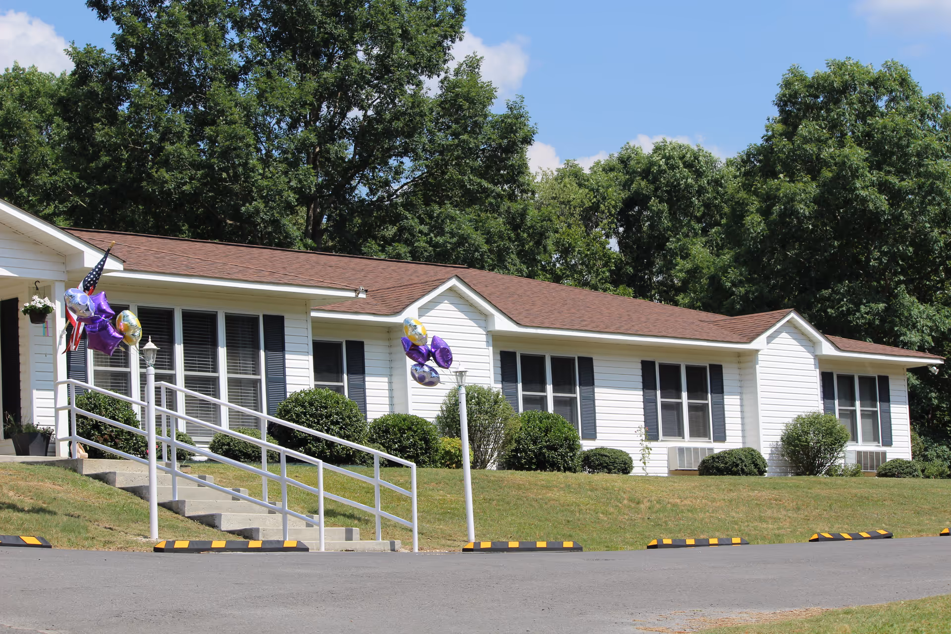Exterior view of a single-story white building with a brown roof, black window shutters, and several bushes in front. There is a concrete ramp with white railings decorated with purple and gold balloons, and an American flag near the entrance. The building is surrounded by green trees and a clear blue sky.