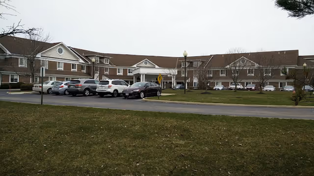 Exterior view of a large two-story senior living facility building with a brown roof and beige walls. Several cars are parked in front of the building on a paved parking lot. There is a grassy area in the foreground with a few small trees and a cloudy sky overhead.