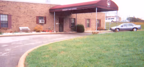 Exterior view of a brick building with a maroon awning labeled 'PERSONAL CARE CENTER' over the entrance. There is a curved driveway in front, a white bench near the building, some bushes, and a parked car on the right side. The foreground shows a grassy area.