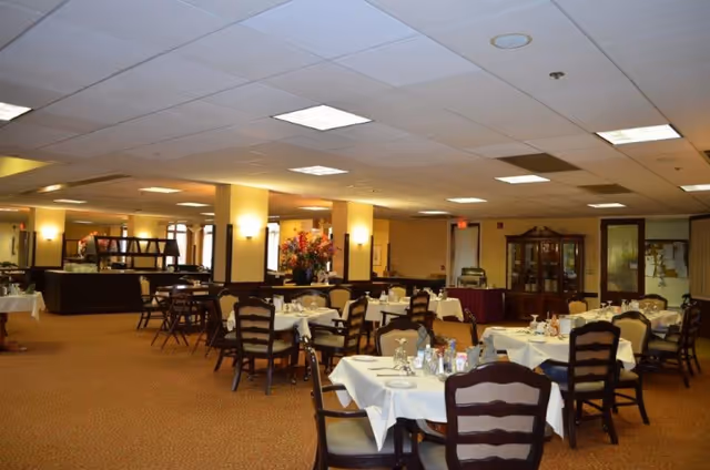 Spacious dining room with multiple round and rectangular tables set for meals, wooden chairs, and a buffet area under warm lighting.