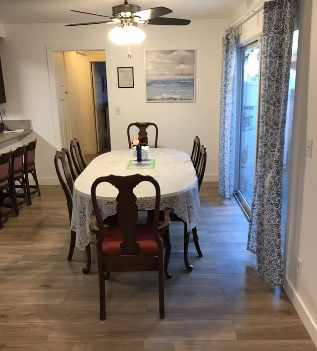 Dining room with an oval table covered by a lace tablecloth, wooden chairs, a ceiling fan, and a sliding glass door with patterned curtains.