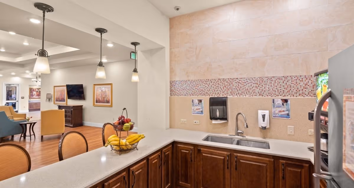 Interior view of a senior living community kitchen area with a countertop, sink, and cabinets. The countertop has a tiered fruit basket with bananas and apples. The kitchen opens into a common area with chairs, a TV mounted on the wall, framed artwork, and wood flooring. Pendant lights hang from the ceiling above the counter.