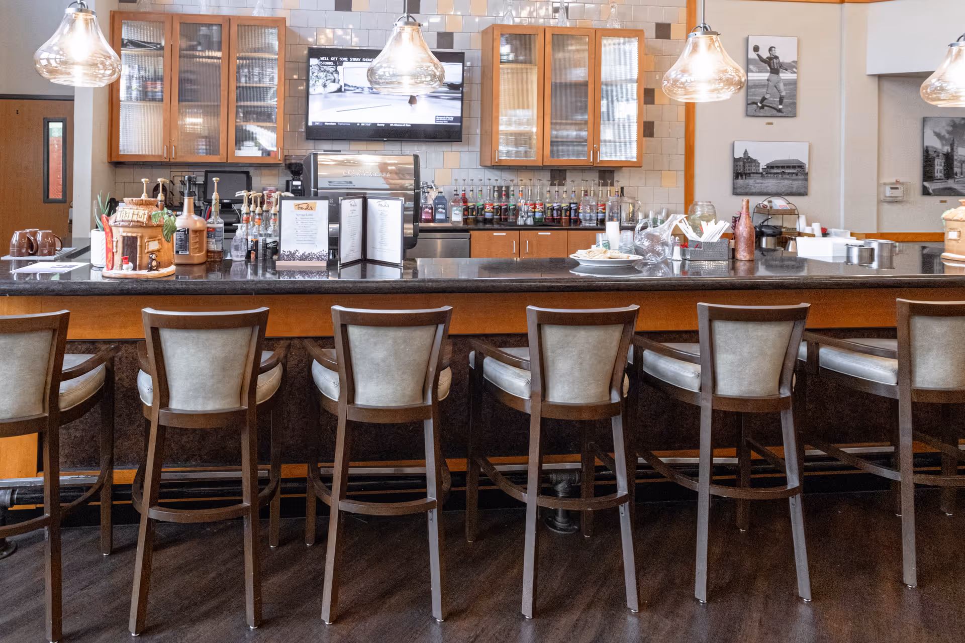 Interior view of a bar area with a row of wooden bar stools with cushioned seats in front of a counter. Behind the counter are shelves with glassware and bottles, a mounted TV showing a basketball game, and pendant lights hanging from the ceiling. The walls have framed black and white photos.