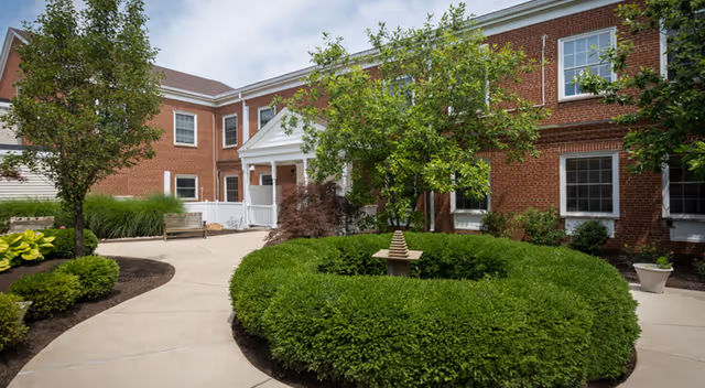 Outdoor courtyard area at Ohio Living Mount Pleasant featuring a circular hedge with a small decorative fountain in the center, surrounded by a paved walkway, trees, shrubs, and a red brick building with white-trimmed windows in the background.