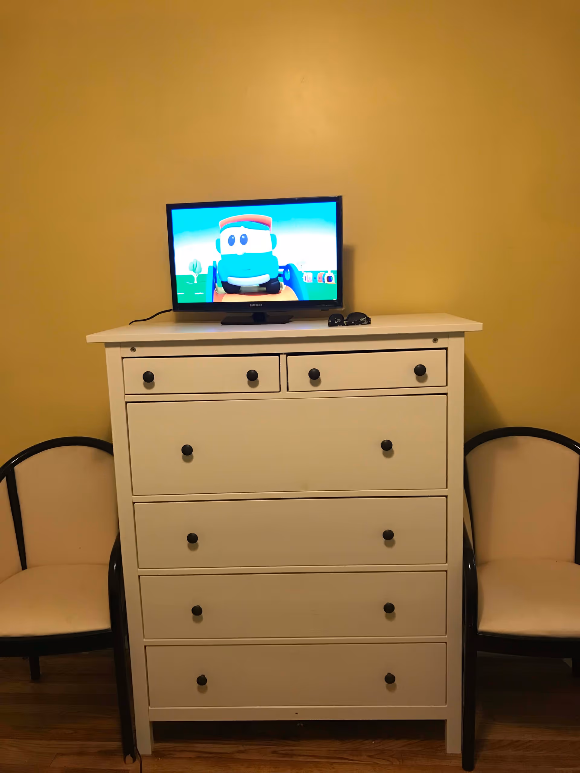 White dresser with a small TV showing a cartoon on top, flanked by two chairs against a yellow wall.