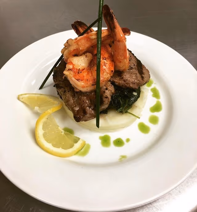 A gourmet dish featuring grilled shrimp placed on top of two pieces of grilled meat, served on a bed of mashed potatoes and sautéed greens, garnished with two lemon wedges and green sauce drops on a white plate.
