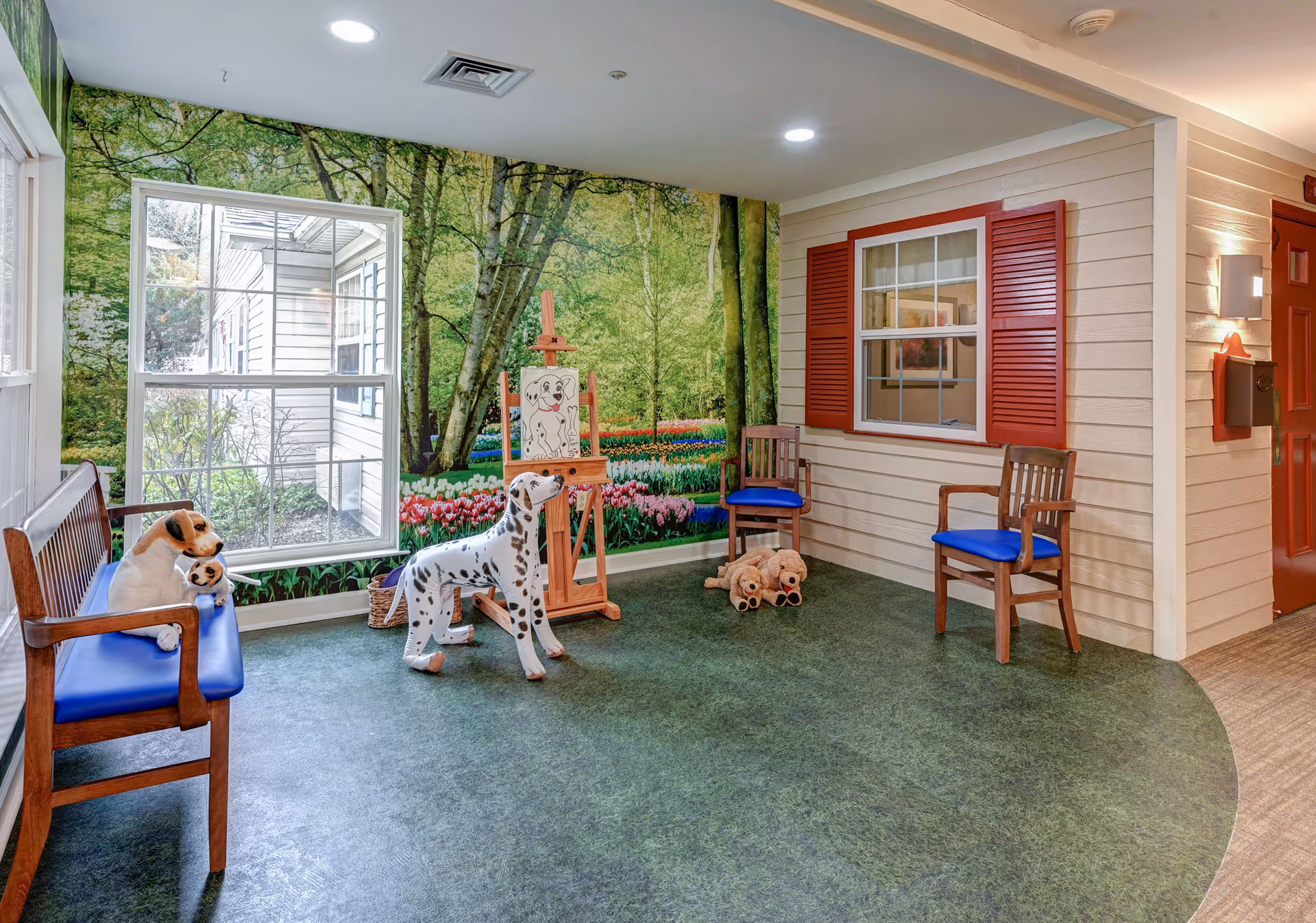 A cozy indoor seating area designed to resemble an outdoor porch with green flooring and a wall mural of a forest and tulip garden. The space includes wooden benches and chairs with blue cushions, stuffed dog toys, a large Dalmatian dog statue, and an easel with a drawing of a dog. There is a window with red shutters and a red door on the right side.