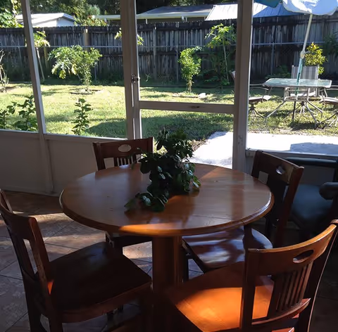 A round wooden table with four wooden chairs around it, placed in a sunlit indoor area with large windows showing a green backyard with grass, small trees, and a patio table with an umbrella.