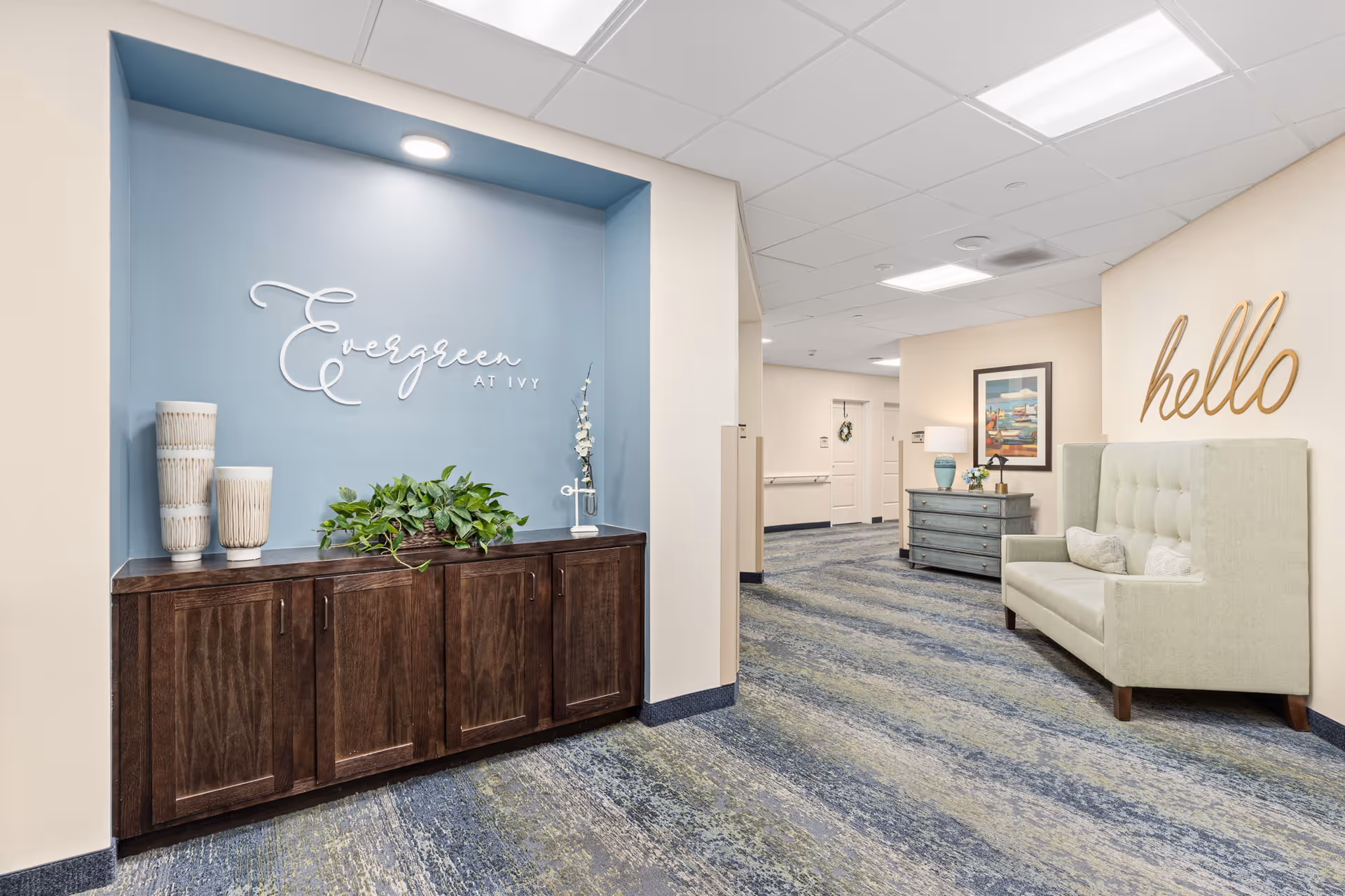 Interior hallway of a senior living facility with a blue accent wall featuring the sign 'Evergreen at Ivy' above a dark wooden cabinet decorated with two vases and a green plant. The hallway has patterned blue and gray carpeting, beige walls, a light green cushioned bench with pillows, a wooden dresser with a lamp and flowers, a framed picture on the wall, and a large wooden 'hello' sign mounted above the bench.