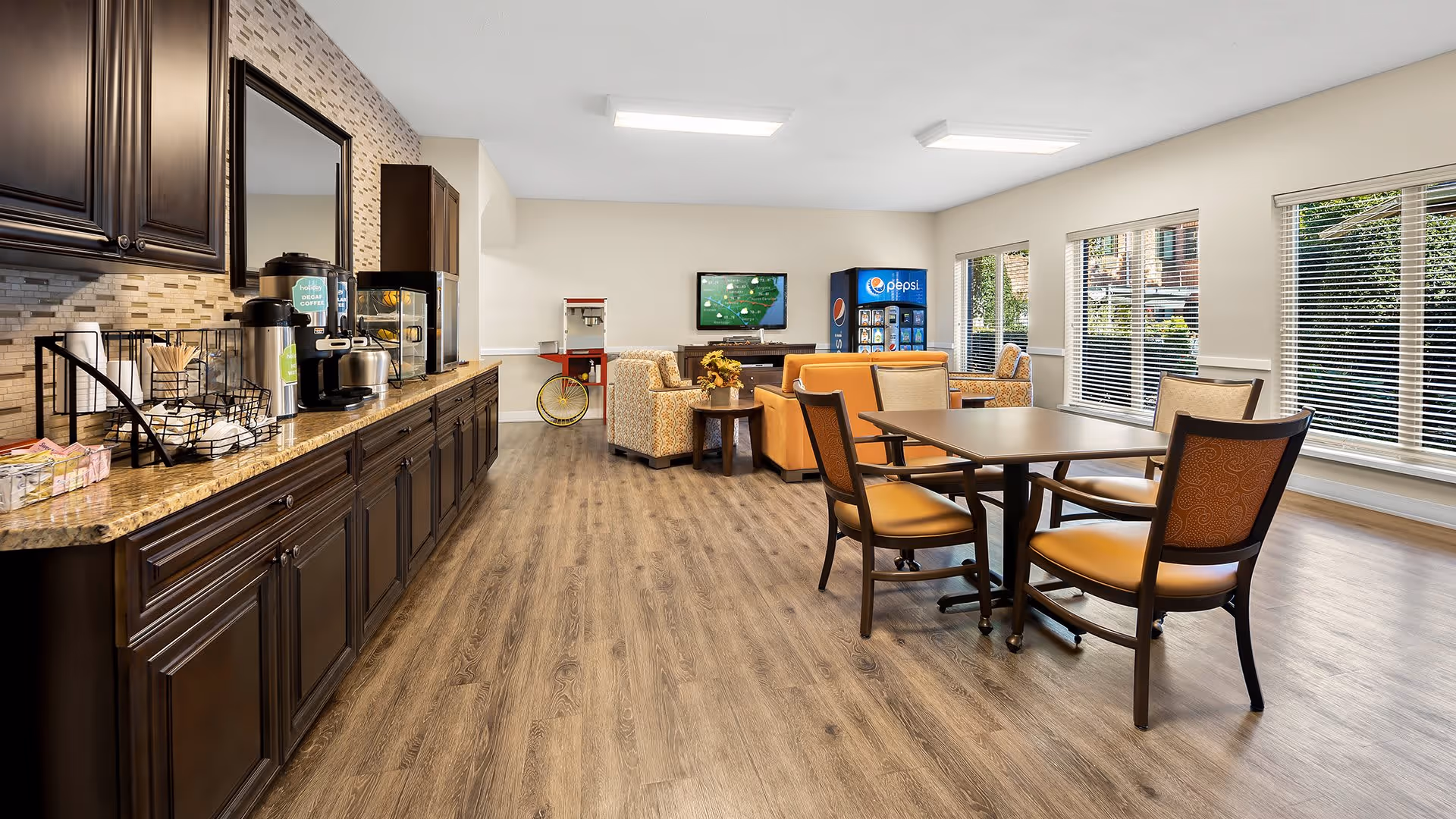 A bright and spacious common area in a senior living facility with large windows letting in natural light. The room features a long counter with dark wooden cabinets, a coffee station, and a snack display. There are several tables and chairs for seating, along with comfortable armchairs and a television mounted on the far wall. A Pepsi vending machine and a popcorn machine are also visible near the seating area.