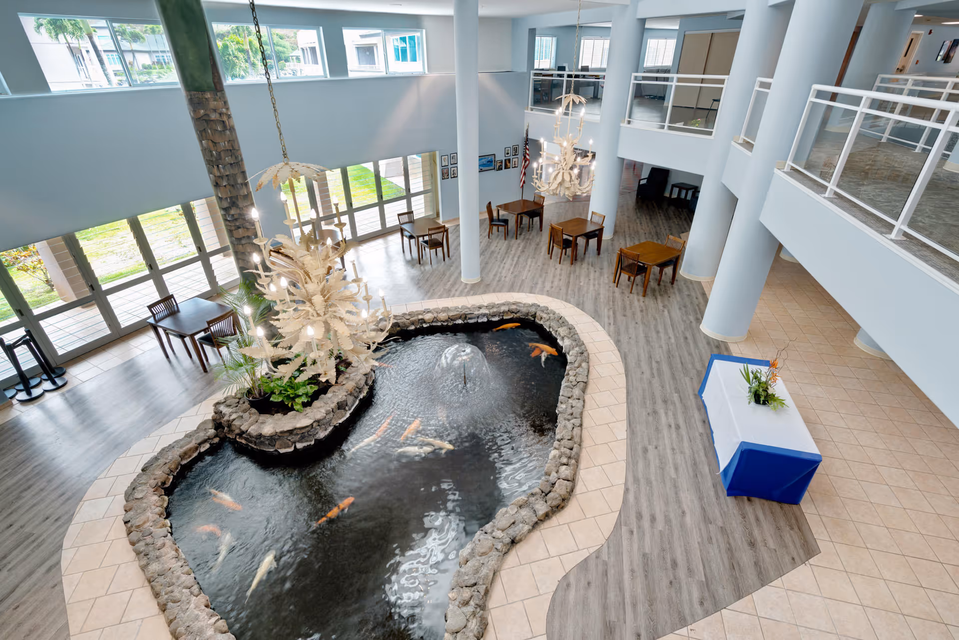 Spacious indoor common area with a large koi pond surrounded by stone, a water fountain in the center, several wooden tables and chairs, large windows letting in natural light, and two white chandeliers hanging from the ceiling.