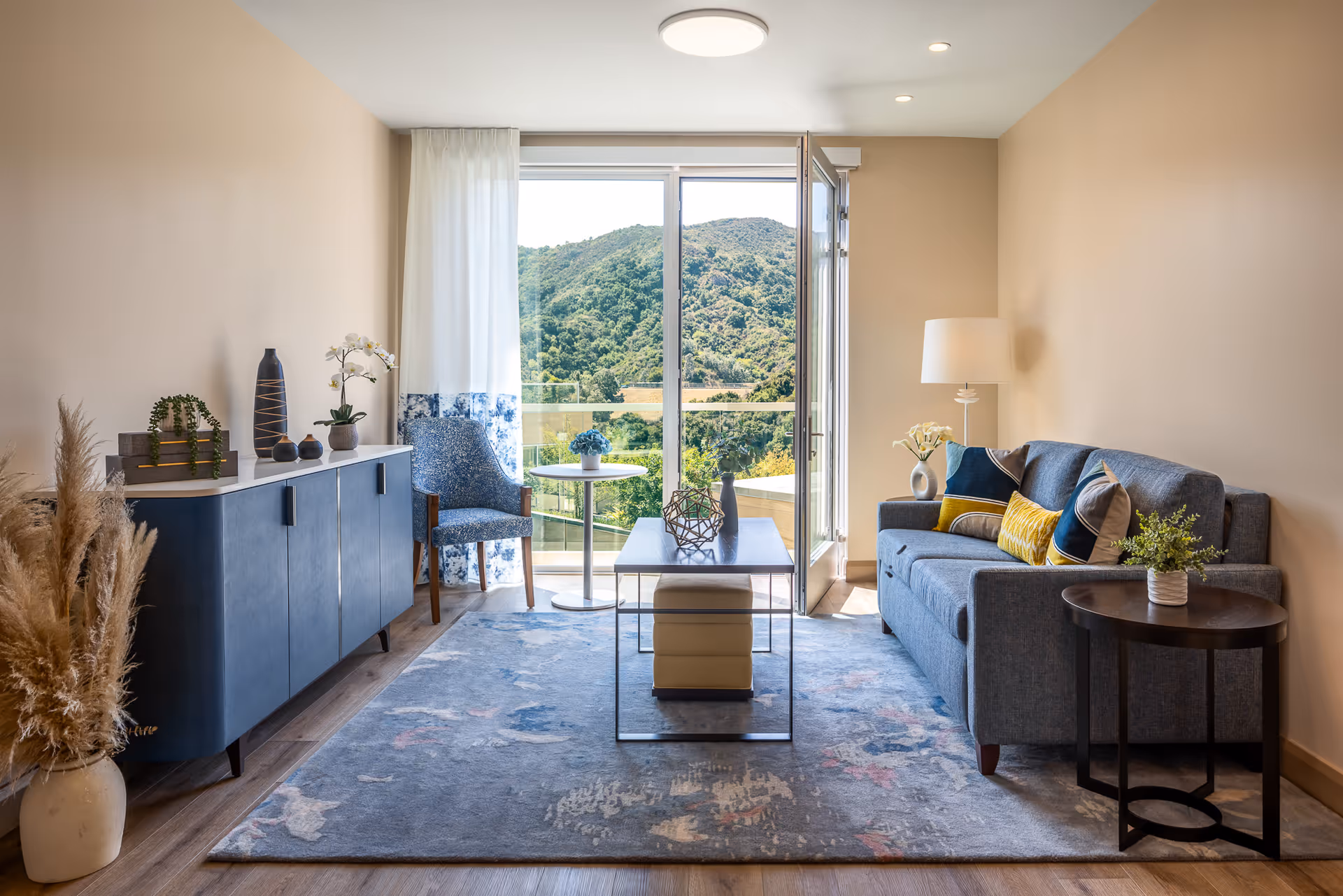 A bright and modern living room with beige walls and wooden flooring. The room features a blue sofa with colorful cushions, a round side table with a plant, a coffee table with decorative items, and a blue cabinet with various vases and plants on top. There is a blue patterned chair next to a small round table by a large glass door that opens to a balcony with a scenic view of green hills and trees. A floor lamp stands beside the sofa, and a large area rug covers the floor.