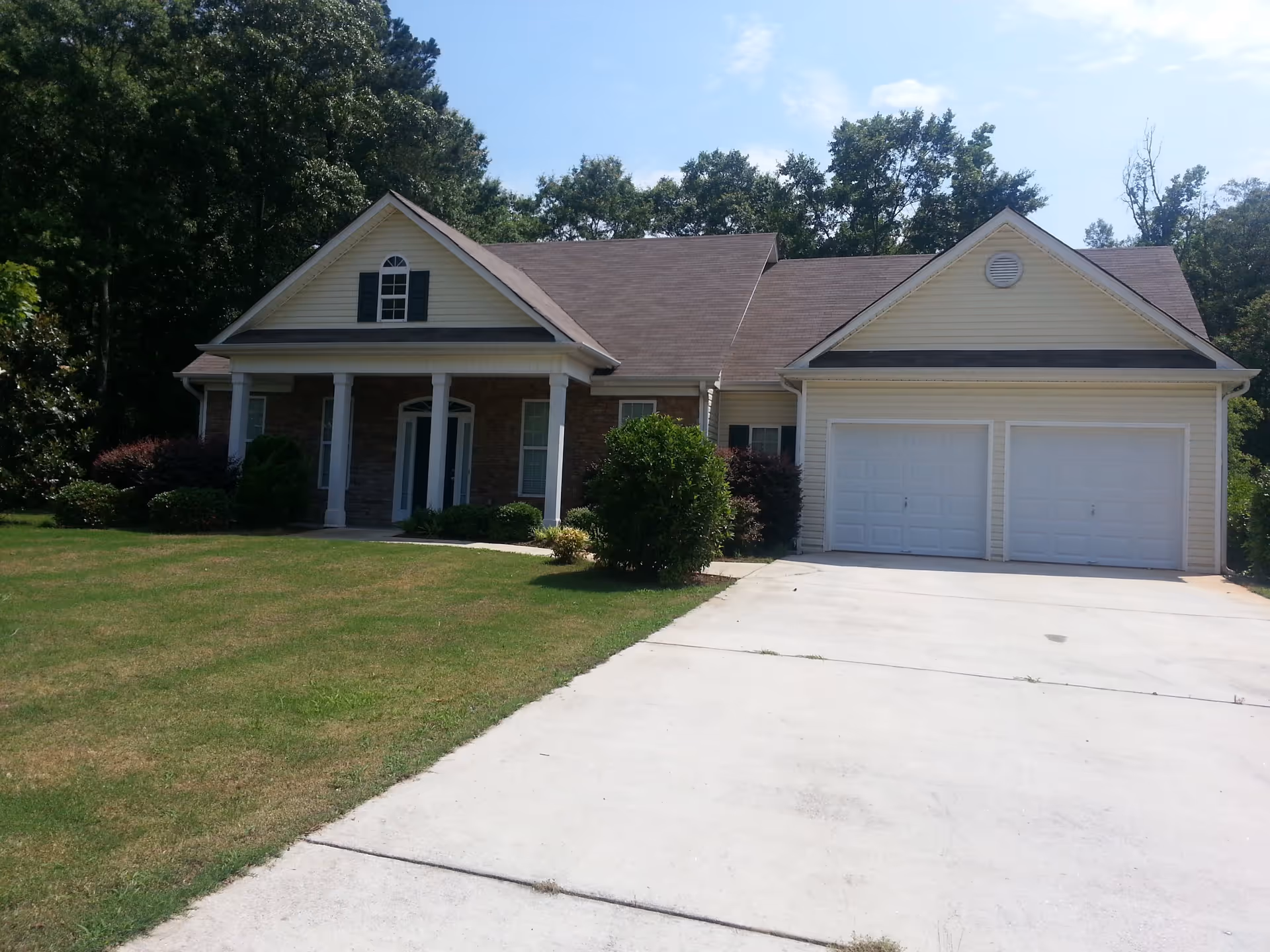 Front exterior view of a single-story house with a double garage, a covered porch with white columns, and a well-maintained lawn with bushes and trees in the background under a clear sky.