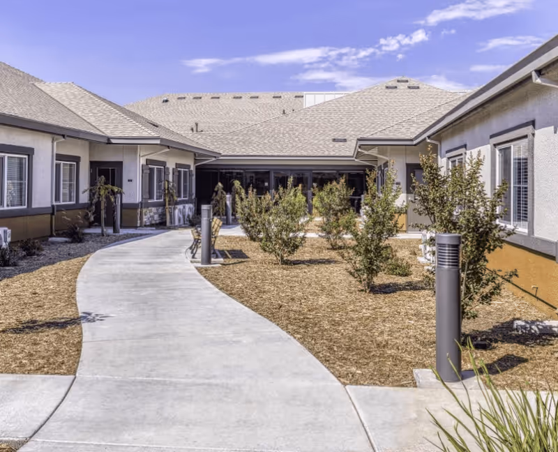Outdoor courtyard area of a senior living facility with a curved concrete walkway, small bushes, and benches. The building surrounds the courtyard with multiple windows and a light-colored roof under a partly cloudy blue sky.