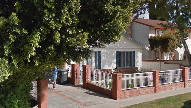 View of a residential-style building with white exterior walls and blue window shutters, surrounded by a low brick and white metal fence. There is a large tree on the left side partially obscuring the view, and a sidewalk in front of the property. A person is standing near a trash bin by the driveway.