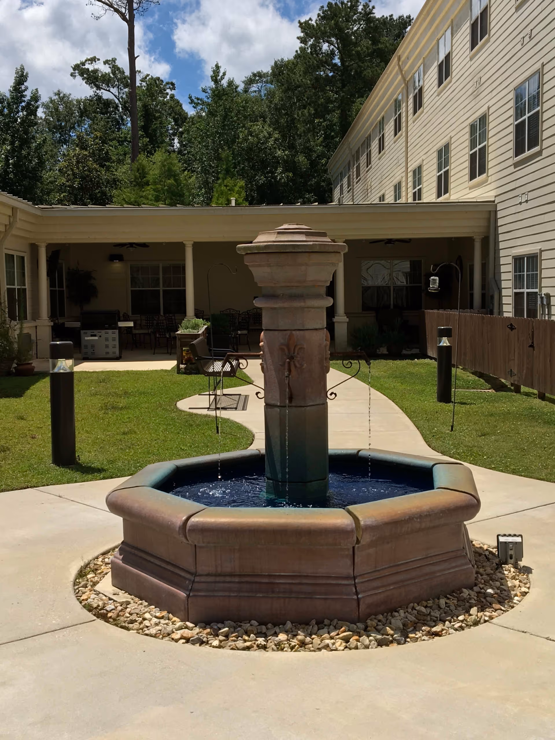 Outdoor courtyard area with a central stone water fountain surrounded by a concrete walkway and green grass. The courtyard is bordered by a multi-story beige building with several windows and a covered patio area with seating and a grill. Trees and a partly cloudy sky are visible in the background.