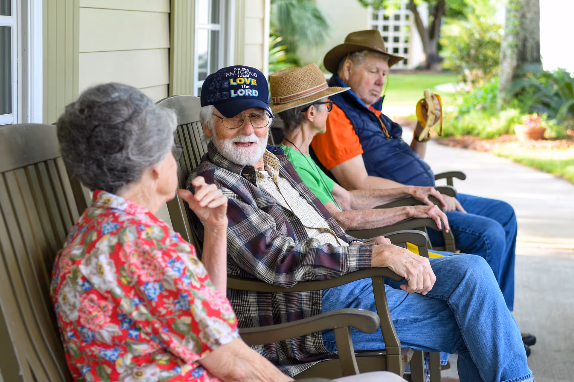 Four elderly people sitting on wooden rocking chairs on a porch outside a building. They are engaged in conversation and relaxing, with greenery and a walkway visible in the background. One man is wearing a hat and holding a banana.