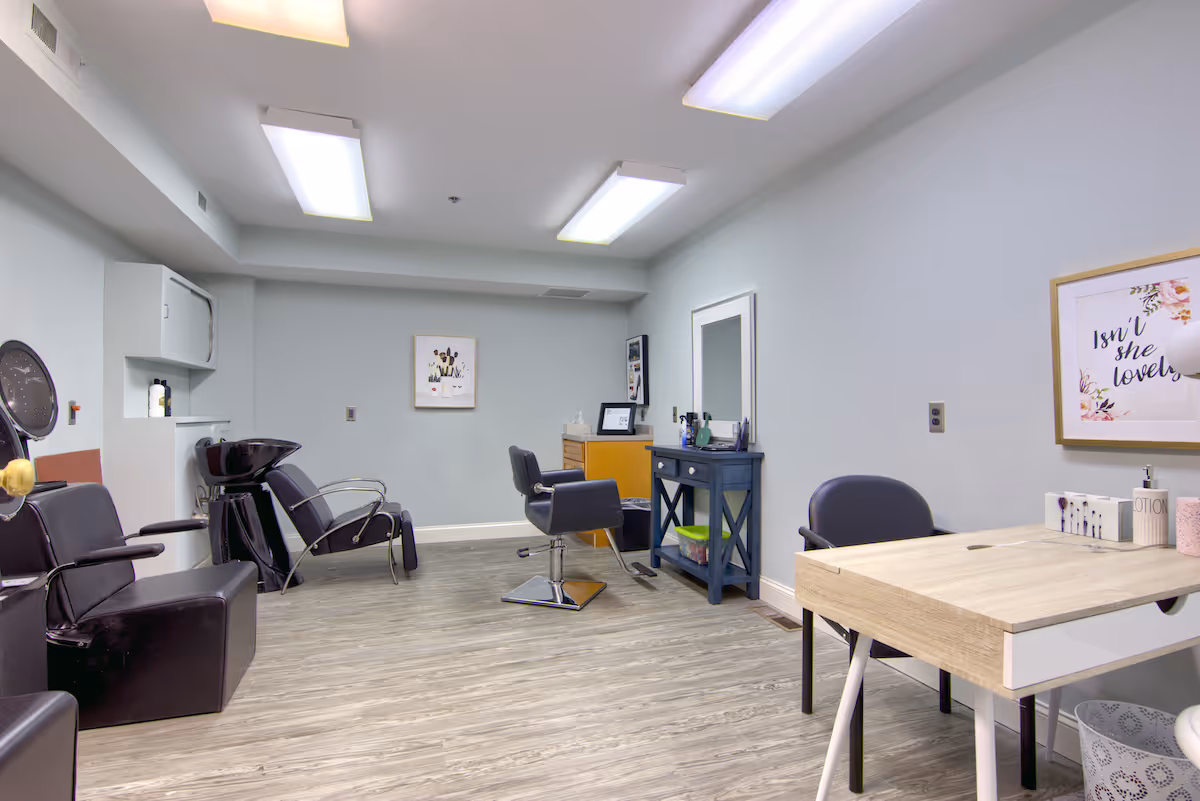 Interior view of a salon area in a senior living community with black salon chairs, a hair washing station, a vanity with a mirror, and a small desk with a chair. The walls are light blue and decorated with framed artwork, and the floor has wood-like vinyl flooring.