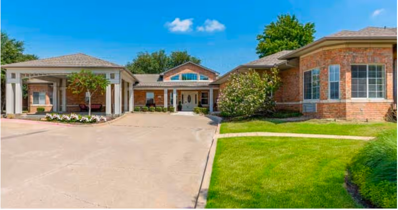 Front exterior view of Spring Creek Assisted Living facility showing a single-story brick building with a covered entrance, well-maintained green lawn, and clear blue sky with a few clouds.