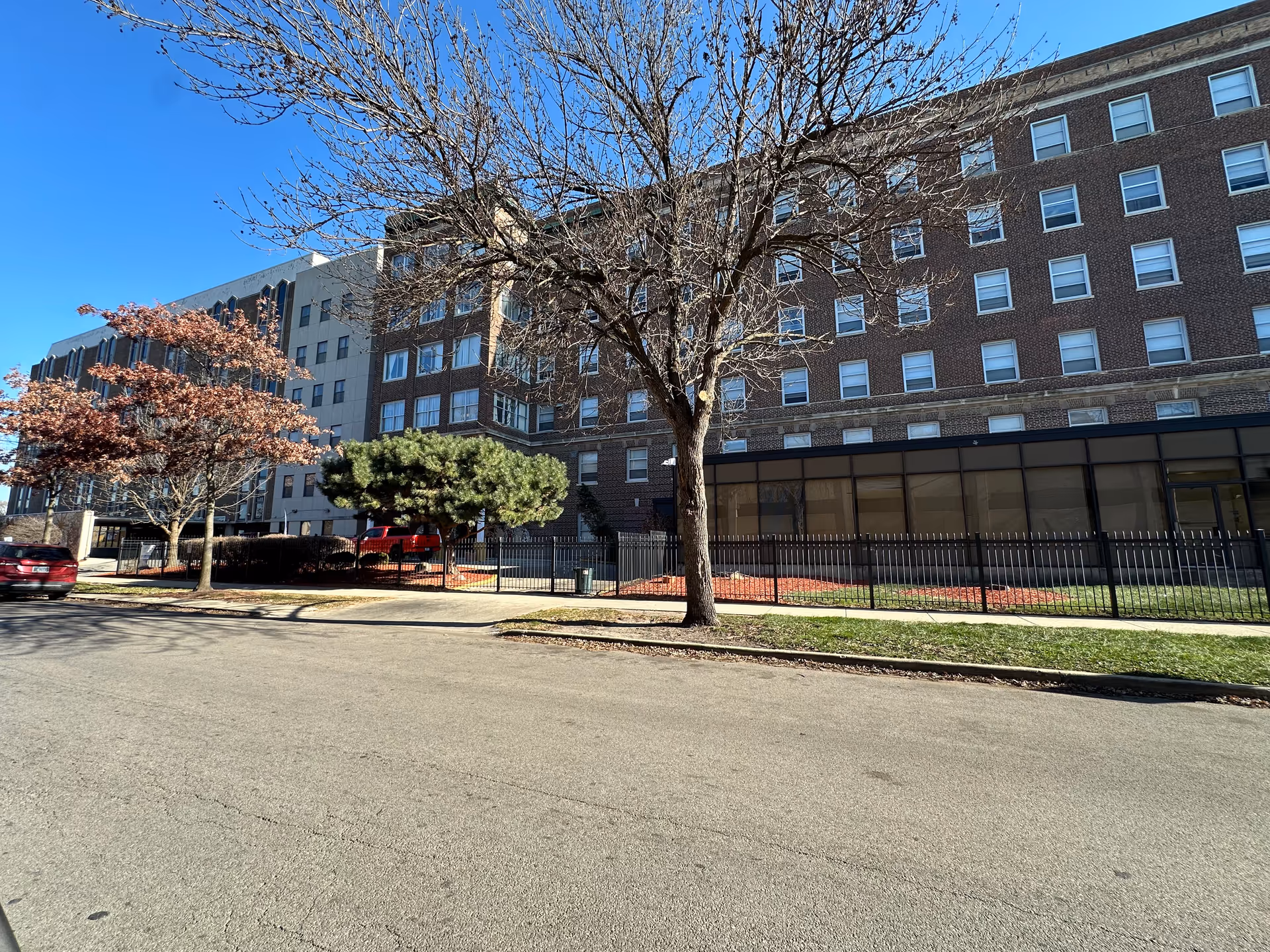 Exterior view of a multi-story brick building with many windows, a fenced area with some trees and shrubs, and a clear blue sky above. There are a few cars parked along the street in front of the building.